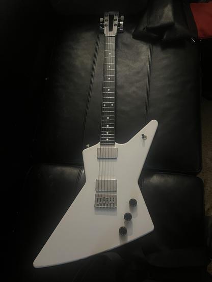 An electric guitar with a distinctive angular design, finished in white, resting on a black leather surface. The guitar features a metal bridge, chrome hardware, and three control knobs.