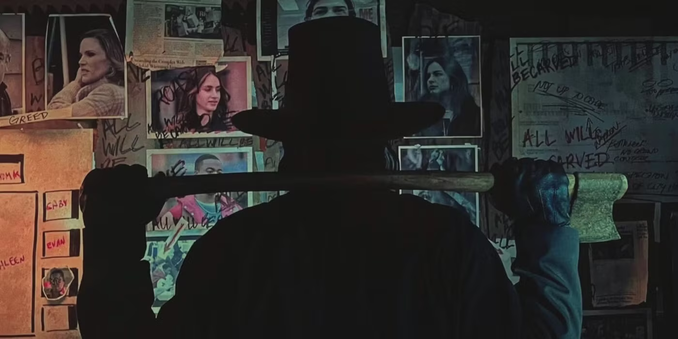 The John Carver villain from the 2023 horror movie Thanksgiving. He is standing, facing away from the camera, staring at a wall of photos with writing on them. He is holding an axe across his shoulders with both hands.
