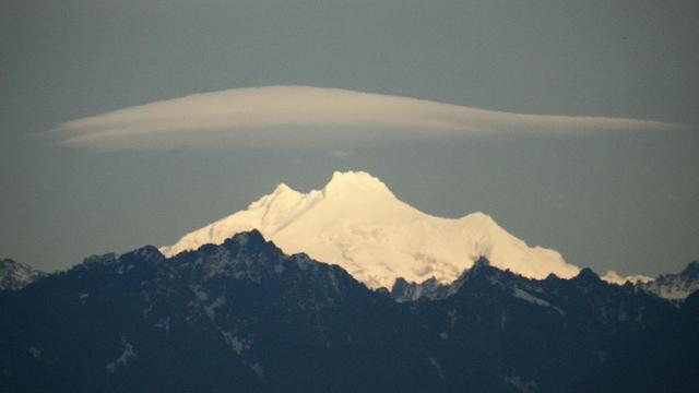 A snow-capped mountain peak partially shrouded by clouds, with a dramatic, single lenticular cloud formation above it, against a grey sky. Surrounding less snow covered mountains are visible in shadow, creating a rugged mountainous landscape.