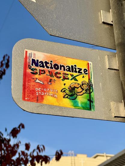 A colorful slap-sticker attached to a metal sign reads "Nationalize SpaceX," with smaller text below saying "de-orbit starlink". A cartoon astronaut floats above the main text. This art was drawn on a USPS Priority Mail label. The sign is attached to a metal post against a clear blue sky.