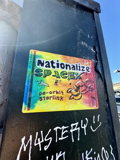 A USPS Priority Mail label that has been dyed bright colors, and had art painted on top depicting Elon Musk in space suit. He is flicking off the viewer, behind him a spaceshuttle and satellite are about to collide. Text on the sticker says "Nationalize SpaceX. De-orbit Starlink.". This label is affixed to a large public utilities box on the street and it is surrounded by other graffiti.