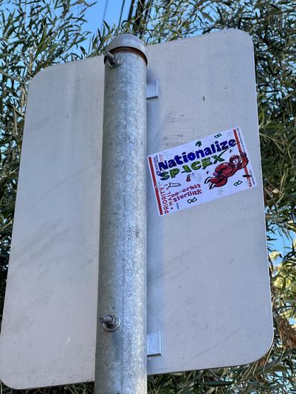Another sticker slapped high up on the back of a parking sign. The sticker says "Nationalize SpaceX, de-orbit starklink". It is drawn on a black USPS Priority Mail sticker label. It also has a drawing of Elon Musk floating in a space suit and flicking off the viewer with his middle finger. 
