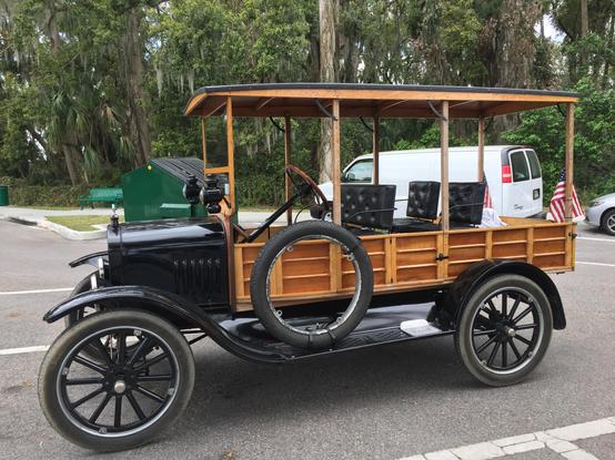 An antique auto, a station wagon with a wood body, the rest of the auto is black.  Probably from the 1920s.  It is sitting in a parking lot with trees in the background.