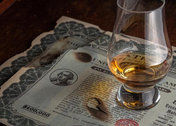 A photograph of a Glencairn glass of whisky sitting on top of some (prop) bearer bonds that are a bit singed around the edges, as if they'd been through, say, an explosion at Nakatomi Plaza. 
