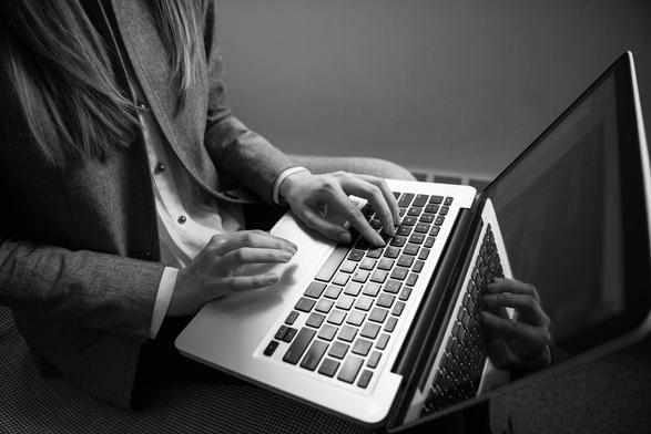 Partial grayscale photo of a woman wearing a suit jacket, seated in front of a laptop with her hands poised over the keyboard