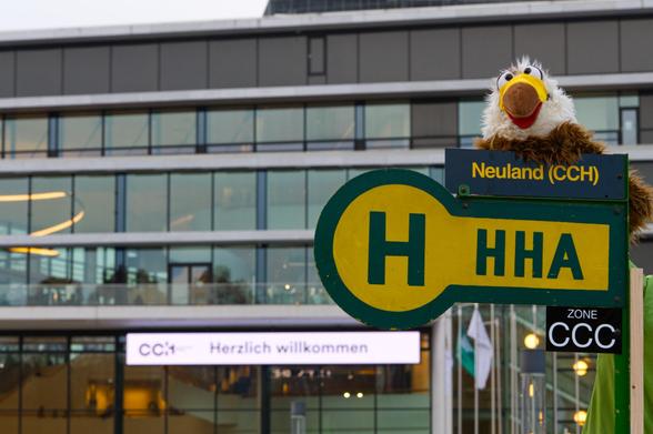 Adelheid sitzt auf einem alten Bushaltestellenschild vor dem CCH. Der Name der Haltestelle ist "Neuland (CCH)", die Tarifzone "CCC". Im Hintergrund das CCH mit dem Schild "Herzlich willkommen".