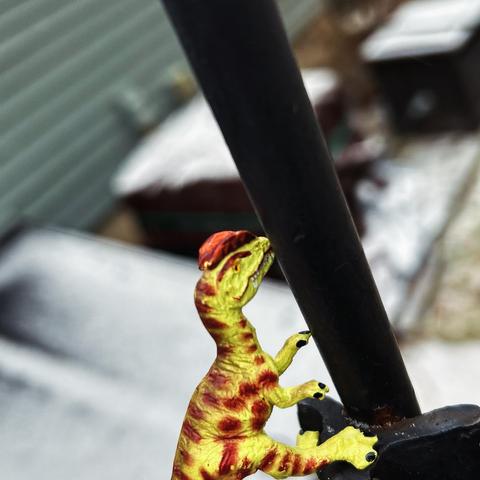 A green Dilophosaurus with brown spots with their tongue stuck to a black metal pole in a snowstorm.