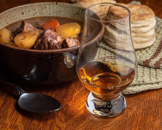 A photograph of a Glencairn glass of whisky next to a fired clay bowl of beef stew on a wooden table, with some flaky homemade biscuits visible in the back of the scene on a pale green knitted tea towel with a brown stripe.  
