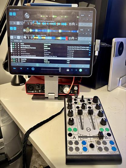 A 12.9” iPad Pro, on a magnetic stand, running Tau DJ software.  A FaderFox DJ2 MIDI DJ controller is in front of it, on the desk.  Behind the iPad is a Focusrite Scarlet 2i4 Gen 2 Audio & MIDI Interface.