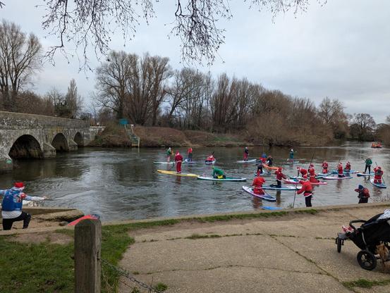 River scene with people dressed as Santa, standing, or sitting on paddle boards, watched by child in a pram.