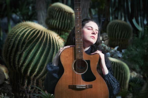 Pressefoto von Reni. Sie hält eine Gitarre vor sich und blickt verträumt nach oben. Im Hintergrund sind große, exotische, runde Kakteen.