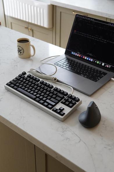 A silver keyboard with black keycaps on a white countertop. It is plugged into a laptop.