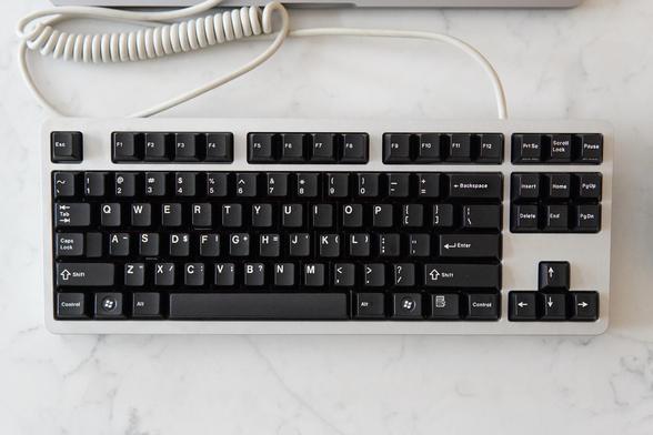 A top down view of a silver keyboard with black keycaps. There is a cream colored cable with a coil connected to it. It’s resting on a white countertop.