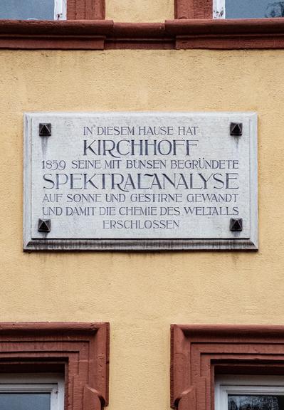 A stone plaque on a yellow wall commemorating the achievements of Gustav Kirchhoff in establishing spectral analysis in 1859. The text is in German and mentions his work with Robert Bunsen on solar and stellar chemistry.