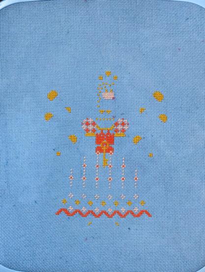 A parially finished cross stitch pience on a medium blue fabric. So far there is some yellow spots floating around, a red border on the bottom, and a pink and red checked pattern in a small area in the middle.