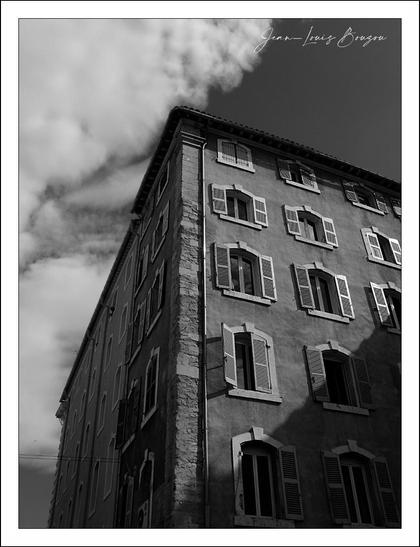 The image is a black-and-white photograph of a tall building, viewed from an upward angle. The building has multiple stories with several windows, each equipped with shutters. The façade is divided by a visible vertical line of stone. There are clouds in the sky, adding depth and contrast to the scene. The photograph captures a sense of architectural elegance with its thoughtful use of light and shadow.
