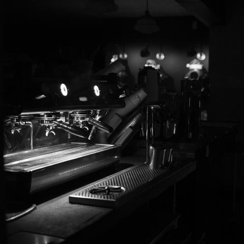 A black and white photo of an unused, but lit espresso machine at night through the window of a closed cafe in Alameda, CA. There are lights visible in the background, but other than that and the light in the coffee preparation area, not much is visible.