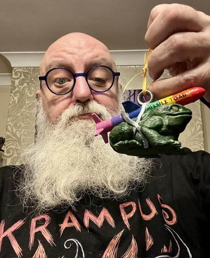 Bespectacled Beardy Weirdy Blok holding an ornament of a toad with a rainbow  missile on its back with the words "Missile Toad" emblazoned upon it. 