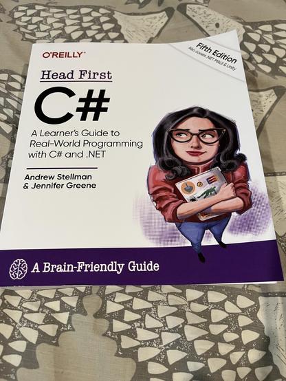 A book "Head First C#, Fifth Edition" sits on a bed, a quilt cover of hedgehogs upon it. 
The grimoire shimmers with promise, its pristine white surface marked with the sigil of O'Reilly, blazoned in the colour of fresh-spilled wine. At its heart stands the rune "C#", bold as a warrior's declaration, flanked by the sacred inscription "A Learner's Guide to Real-World Programming with C# and .NET". The scribes who penned this codex – Andrew Stellman and Jennifer Greene – have etched their names beneath, testament to their mastery of the digital arts.


An illustrated guardian brings this book to life, as they stand watch upon its surface – an enchanting figure with raven-dark waves of hair & eyes that sparkle with wisdom behind crystalline spectacles. Clad in a garment the colour of autumn leaves, she cradles a laptop adorned with mystical sigils, her face in awe of the secrets yet untold. She is both teacher & guide, keeper of coding mysteries, ready to lead worthy students through the labyrinth of programming knowledge.


At the base, a purple banner unfurls like a royal decree, bearing both the symbol of consciousness – a simplified brain sigil & the promise of "A Brain-Friendly Guide". This text rests upon its hedgehog-guarded altar, each quilted spine in the fabric below standing ready to defend its technological wisdom, creating a perfect union of modern sorcery & timeless protection.