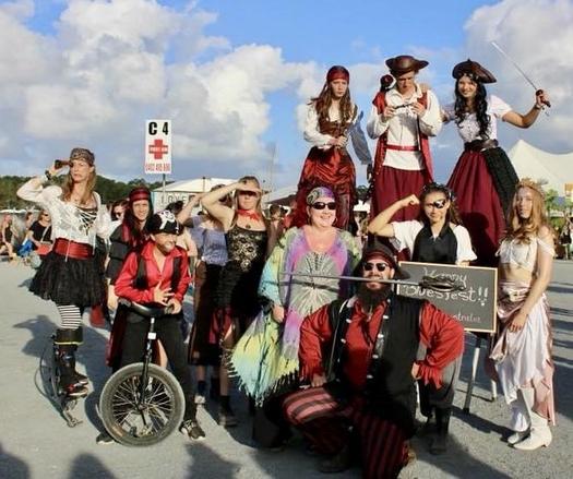 A gathering of pirates stands frozen in time against a sky painted with billowing clouds, their chosen battlefield, the soon to be no more: Byron Bay Bluesfest. Their crew, a vibrant assembly of rogues and adventurers (circus performers and the author), forms a tableau worthy of legend. 

Three bold souls perch atop wooden stilts, their crimson-clad forms towering above their compatriots like the masts of a proud galleon. Below, their fellow buccaneers stand united, each a character sprung from tales told in dimly lit taverns - one astride a mechanical steed of a singular wheel, others adorned in the finest piratical regalia of stripes and ruffles. 

A fairy-like figure sparkles in their midst, her kaleidoscope garments shimmering like captured starlight. 

At the scene's edge, a warrior-scribe ( yep, it me!) brandishes not just blade but body as well, teeth clenched around steel (LARP) in defiance of convention, epitomizing the unorthodox spirit of this unconventional crew. 

Which makes sense because a pirate and tutor of STEM I.e. calculus - we never forget the C(#)!
