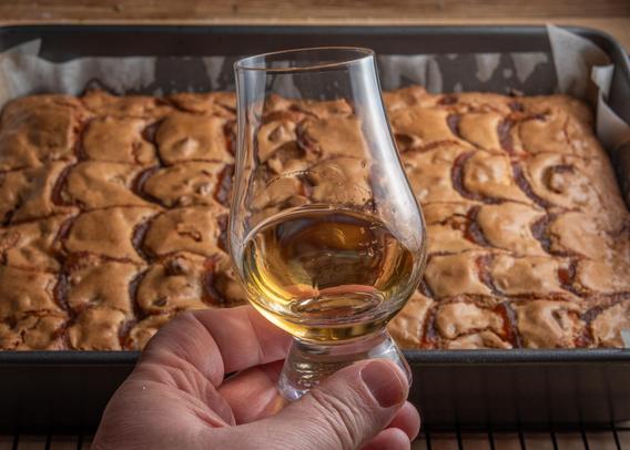 A photograph of a Glencairn glass of whisky being held in the photographer's hand, with a tray of blondies streaked with apple butter in a pan in the background.
