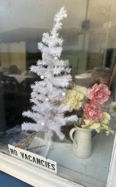 A bed and breakfast dining room, void of people, with a sparsely decorated white Christmas tree and a sign saying “NO VACANCIES”. A jug of fabric flowers sit to the right.