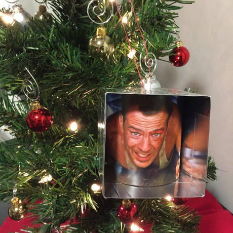 **TL;DR:** A Christmas tree ornament features Bruce Willis as John McClane in the iconic air vent scene from *Die Hard*.
The image shows a decorated Christmas tree with red and gold baubles and lights. Hanging from the tree is a unique ornament shaped like a small box with a scene from *Die Hard* printed on it. The image on the ornament shows Bruce Willis as John McClane crawling through an air duct, a memorable moment from the film. This ornament playfully ties the movie to Christmas, as *Die Hard* is often humorously debated as a Christmas movie.