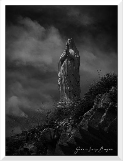 This black and white image depicts a statue of a woman standing atop a rocky hill. The statue appears to be of a religious nature, with the figure in a traditional praying pose, hands together and eyes gently closed. The sky in the background is filled with dramatic clouds, casting a somber and atmospheric mood over the scene. The combination of light and shadow enhances the texture of the statue and the surrounding rocks, giving the image a timeless and reverent feel.
