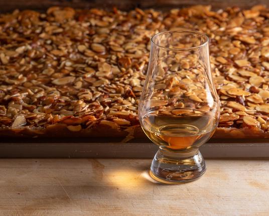 A photograph of a Glencairn glass of whisky with a slab of Florentine cookies visible behind (almonds and candied orange peel in a caramel base on top of shortbread). The cookies are sitting on top of a white cutting board, and everthing is sitting on a butcher block surface. 

