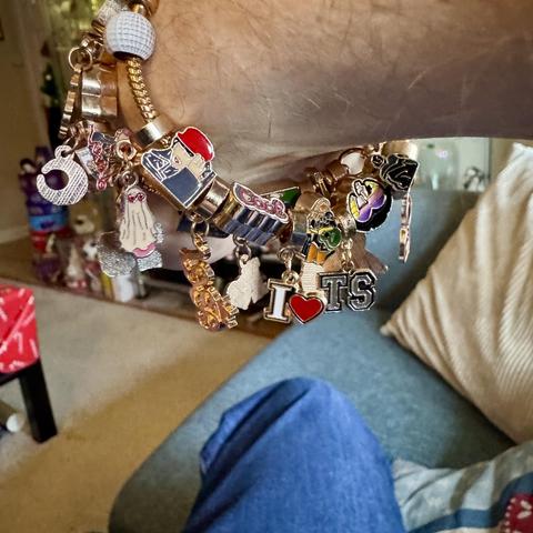 A close-up of a wrist adorned with a charm bracelet featuring various colorful charms, including illustrative images of Taylor Swift like the cover of Red (Taylor’s Version) and text that says things like "I ❤️ TS" (split into two charms) and “cool”.  The background shows a cozy living space with a sofa and decorative pillows.