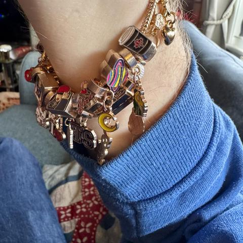 A close-up of a wrist adorned with a colorful charm bracelet featuring various small charms, including letters & symbols spelling out “I❤️ TS,” illustrative images of Taylor Swift, and playful references to her lyrics like a Coffee Cup, a moon & Saturn. The bracelet is layered over a blue sweater.