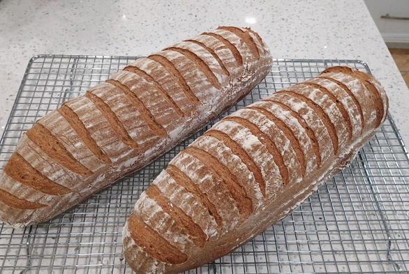 Two loaves of rye bread just fresh out of the oven cooling on a rack. They're long loaves with multiple slashes across the loaf