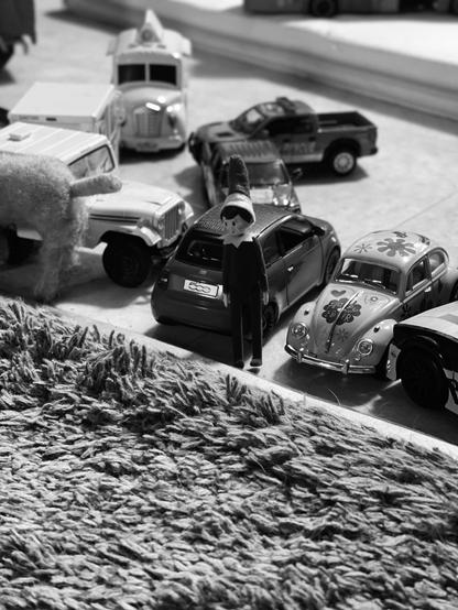 A collection of toy cars and figures on a textured rug, with a prominently featured doll dressed in black and white, resembling an elf. The scene is captured in black and white, highlighting the various vehicles and the doll amidst them.
