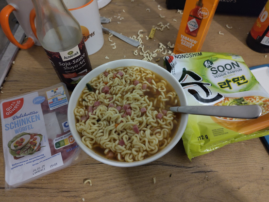 **TL;DR:** A bowl of instant veggie ramen with added pork cubes, surrounded by cooking ingredients and packaging on a cluttered table.  

The image shows a bowl of ramen soup made with "Soon Veggie Ramyun Noodle" by Nongshim, visible from the packaging nearby. The soup is topped with diced ham or pork cubes from a pack labeled "Gut & Günstig Delikatess Schinken Würfel." A bottle of soy sauce (Alnatura Soja-Sauce Shoyu-Art) and orange Tabasco sauce are placed nearby, along with an orange mug and loose noodles scattered around the wooden table. The scene appears casual and improvised, reflecting a humble and solitary Christmas meal setup.