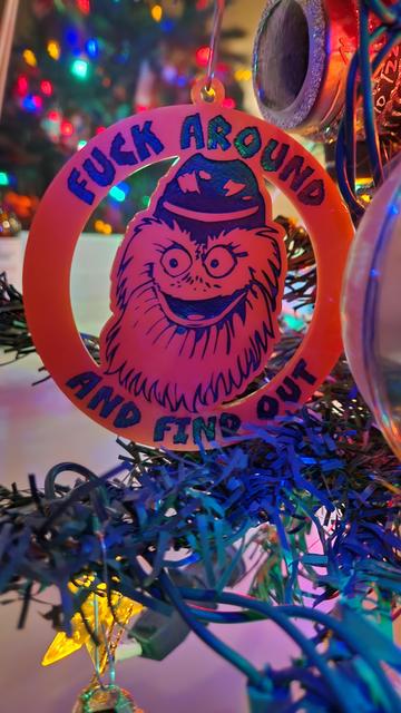 A disc-shaped orange plastic Christmas tree ornament featuring an etched and inked portrait of the mascot Gritty. Gritty is smiling maniacally. The outer ring of the disc reads FUCK AROUND above the portrait of Gritty's head, AND FIND OUT below. The lights of the Christmas tree are reflected in a nearby window.