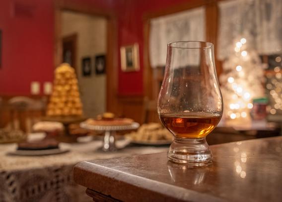 A photograph of a Glencairn glass of whisky on a pink marble surface, with a view to a dining room with a table loaded with baked goods and a small white Christmas tree lit up with a string of lights visible in the background.

