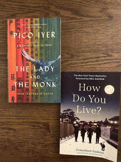 An overhead view of two books on a wooden surface. The top book is "The Lady and the Monk: Four Seasons in Kyoto" by Pico Iyer, featuring a colorful cover with a woman's hand against traditional Japanese letters. The bottom book is "How do you Live" by Genzaboro Yoshino, featuring a dark colored cover with four young men walking at night from a distance.