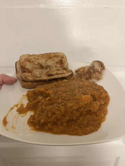 A plate featuring a large serving of Pav bhaji, accompanied by two slices of toasted bread and a small piece of crust. The meal is presented in a hand-held position against a plain background.