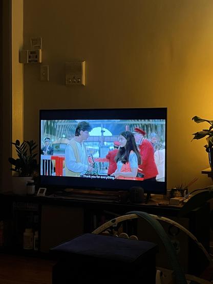 A scene from a movie is displayed on a television, featuring two characters in conversation, with a subtitle that reads "Thank you for everything." The background shows an train station with additional characters in colorful outfits. Nearby, a shelf with plants and various items