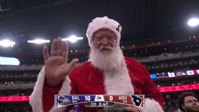 A person dressed as Santa Claus waves, wearing a red suit and white beard, in a stadium during a football game. The scoreboard shows the Baltimore Ravens leading the Houston Texans 10 to 0.