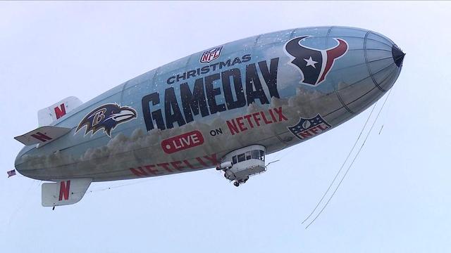 A large blimp featuring NFL branding, including the Baltimore Ravens and Houston Texans logos, promotes a Christmas-themed event on Netflix. The blimp is floating in a cloudy sky with the text "Christmas Gameday Live on Netflix" prominently displayed.