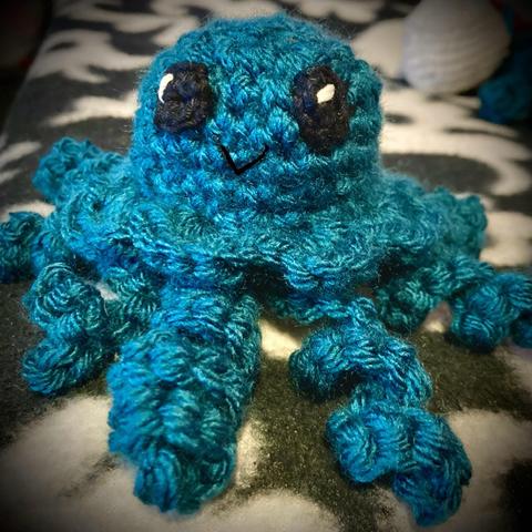 A crocheted jellyfish toy in shades of teal, with large black eyes and a small smiling mouth, sitting against a patterned background, with splayed tentacles.