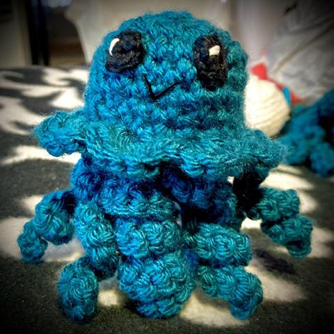 A crocheted jellyfish toy in shades of teal, with large black eyes and a small smiling mouth, sitting against a patterned background.