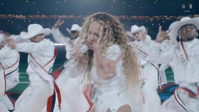 A performer with curly hair (Beyonce) sings into a microphone on stage, surrounded by dancers in white outfits and hats. The background features stadium lighting, suggesting a live performance setting.
