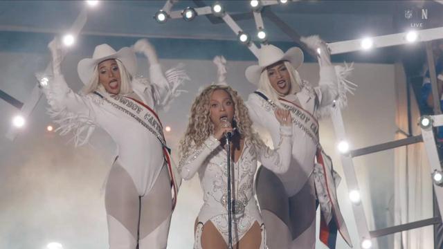 A female performer with curly hair (Beyonce) sings into a microphone while wearing a sparkly white outfit. She is flanked by two backup dancers, also in white outfits with cowboy hats, who are energetically posing. The background features stage lights and a lively performance