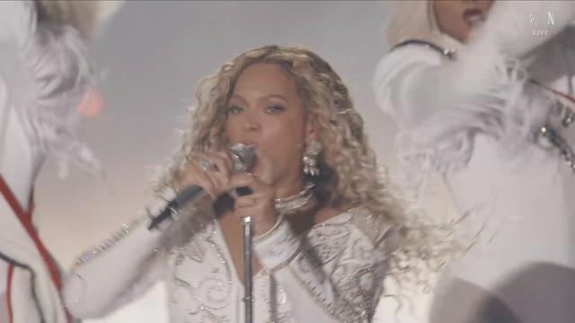 A performer with long curly hair (Beyonce) is singing into a microphone, wearing a sparkling white outfit with intricate designs. Background dancers are partially visible, adding to the dynamic stage presence.