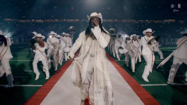 A performer in a white outfit and cowboy hat (Shaboozey)is center stage, singing into a microphone. Surrounding them are musicians in white uniforms, holding instruments and marching on a red carpet laid out on a football field, with stadium lights and smoke in the background