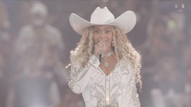 A singer wearing a white embellished outfit and a wide-brimmed white hat (Beyonce) performs on stage, holding a microphone and smiling at the audience.