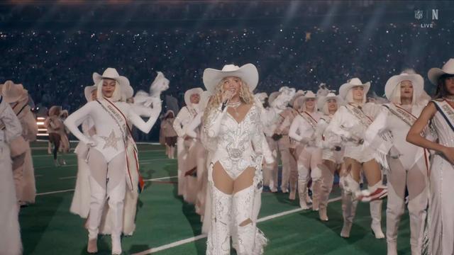 The image shows a performer in a sparkling white outfit and a wide-brimmed hat singing on a football field (Beyonce), accompanied by a group of dancers dressed in similar outfits. The audience in the background is illuminated by lights.