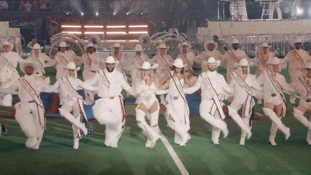 A group of performers in white outfits and cowboy hats dance together on a football field, featuring sparkling sashes and coordinated choreography.